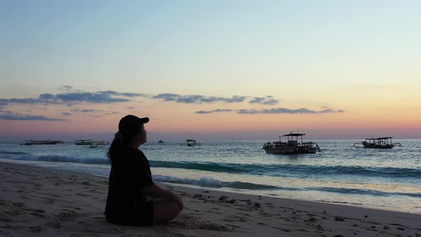 Sporty woman with a hat meditation in the early morning on the peaceful sandy beach. Boats waving on