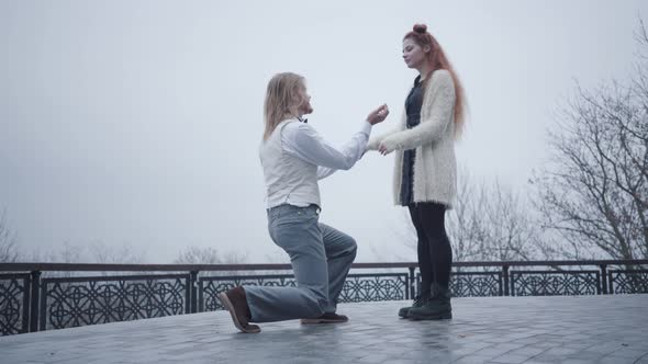 Side View of Stylish Boy in White Shirt and Vest Standing on One Knee and Proposing To His