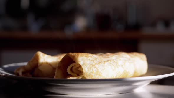 Rolled Up Fried Ruddy Pancakes on a Plate with Rising Steam in Rays of Sunlight