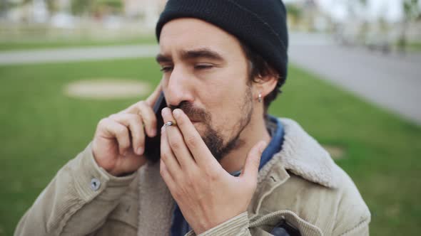 Serious bearded man wearing hat talking by phone and smoking