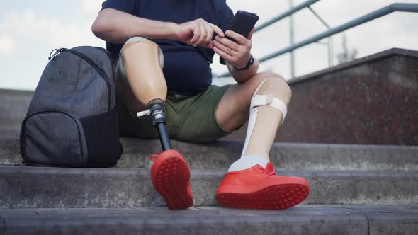 Unrecognizable Man with Prosthetic Legs Sitting on Urban City Stairs Messaging Online on Smartphone