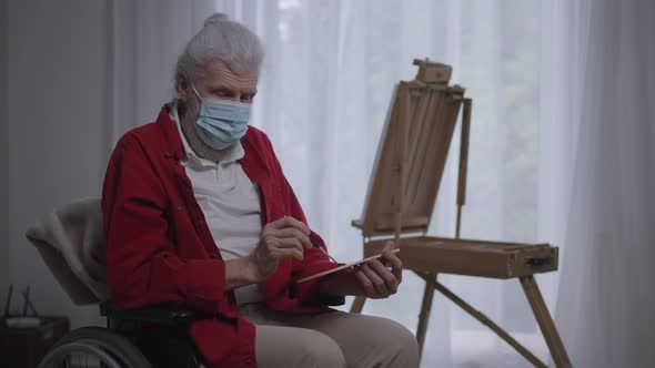 Handicapped Caucasian Old Man in Coronavirus Face Mask Holding Painting Palette and Brush Looking at