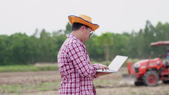 Asian farmer standing and working on laptop at farm and tractor harrowing in background