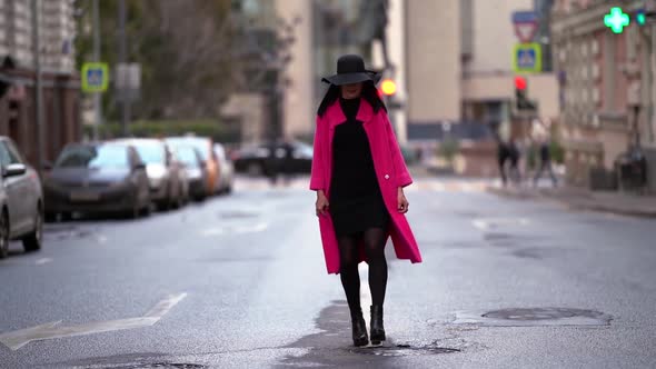 A Beautiful Brunette Woman in a Pink Coat and Black Hat Walks Along an Asphalt Road on a Blurred