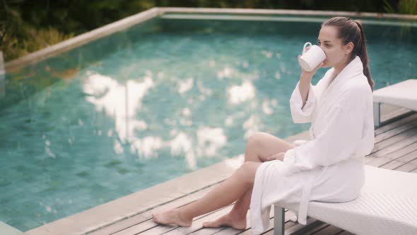 Woman in White Bathrobe Sits on Sunbed Near Pool, Drinks Coffee in Early Morning
