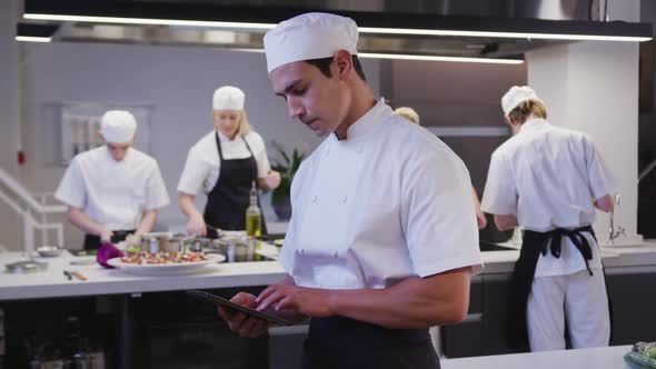 Mixed race male chef wearing chefs whites in a restaurant kitchen using a tablet and looking at came