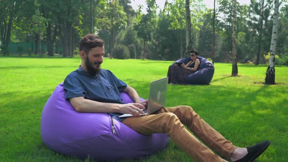 A Young Man in Blue Shirt Sitting in a Chair Ottoman Park and Working on the Computer