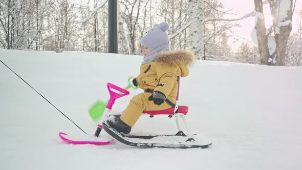 A kid rides a snowcat sled with a shovel toy in his hand along a snowy path in a city park.