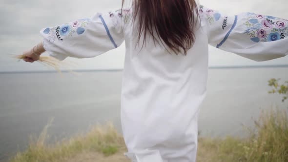 Brunette Girl in Long White Summer Dress with Embroidery Running Among High Grass