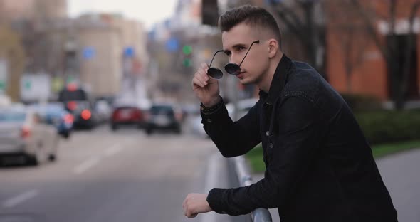 Young Man Leaning on Barriers Looks at the City Car Traffic