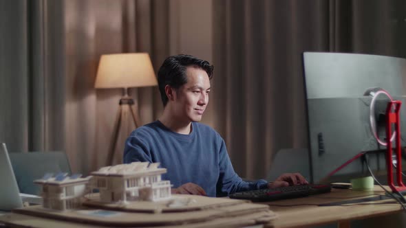 Asian Male Engineer With The House Model Positively Shaking His Head While Working On A Desktop