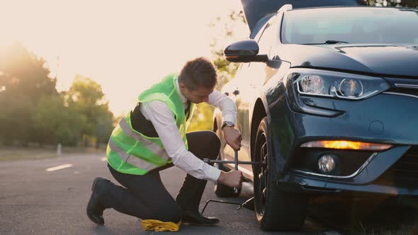 A Man in a Green Safety Vest Changes a Flat Tire on a Road