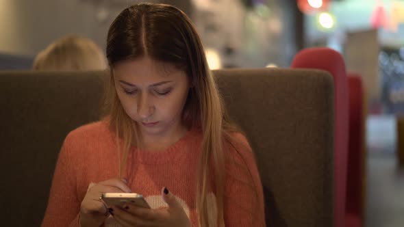 Young Beautiful Girl Sits in a Cafe Works on a Smartphone