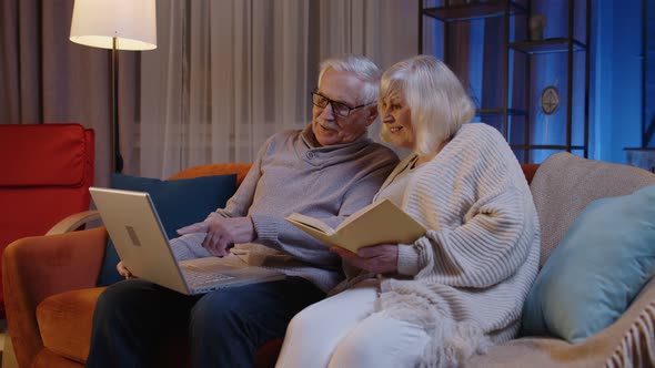 Senior Grandparents Couple Reading Book Using Laptop Pc on Couch in Night Living Room at Home