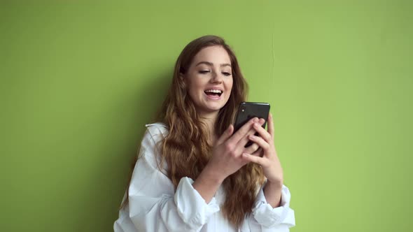 Girl With Long Hair Stands Against Light Green Wall With Mobile Phone In Her Hands.