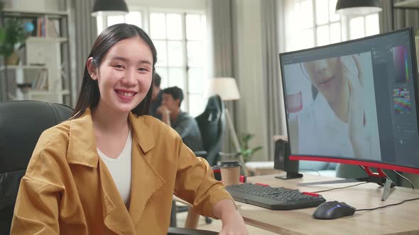 Asian Female Editor Works In Photo Editing Software On Computer. She Turns And Warmly Smiles