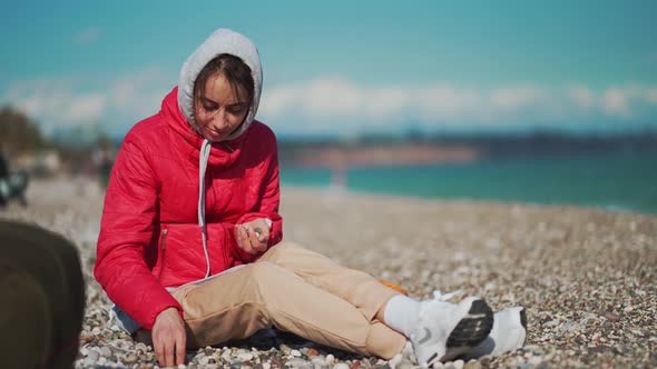 Young Woman in Red Jacket and Hoodie Sitting on Pebble Sea Beach at Windy Sunny Day and Picking Up