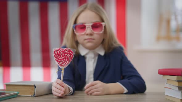 Heart Shape Lollipop in Hand of Joyful Blurred Schoolgirl Sitting at Desk in Classroom