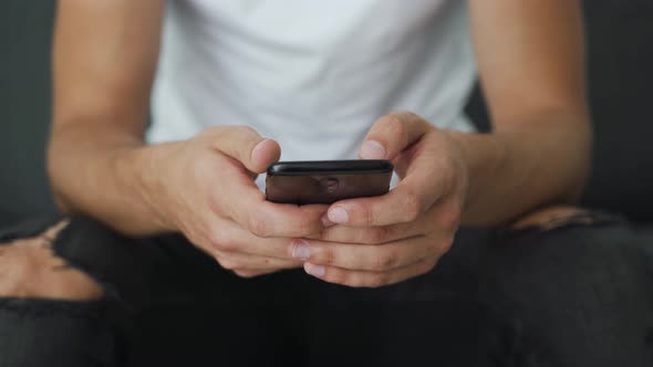 Close up of male teenager hands texting on smartphone while sitting on the sofa. Browse online inter