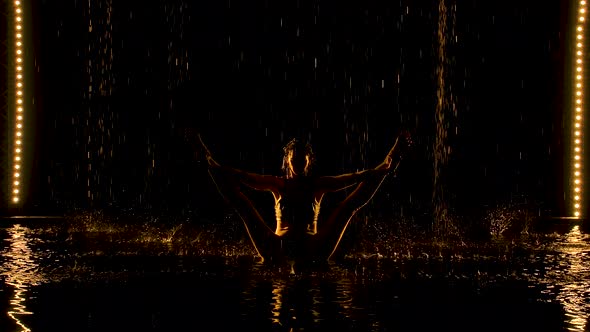 A Young Woman Balance with Legs Wide Apart in a Dark Studio Under the Rain. A Slim Body Silhouetted