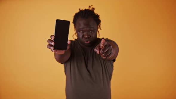 Angry African Man Holding Smartphone and Pointing Fingers Towards Camera