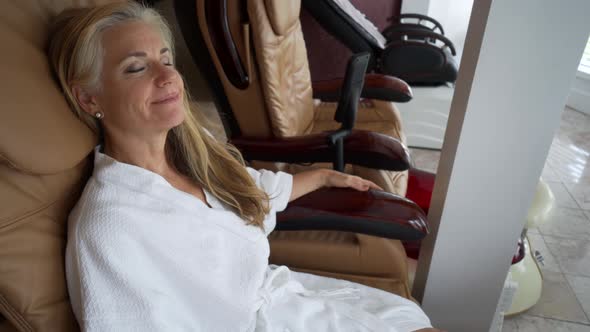 Wearing a white robe, a mature, caucasian woman relaxes back into the thick padding of the pedicure