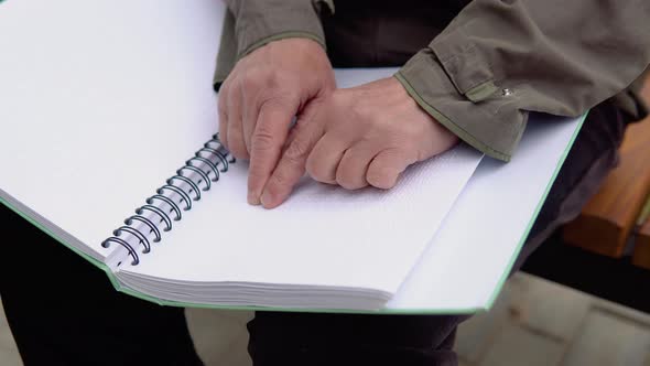 Senior Blind Man Sitting on Bench in City Park and Reading a Braille Book