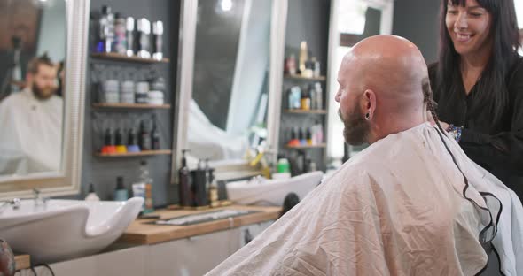 A Female Barber Turned to Camera Brushes Bearded Man's Smoothly Shaved Head Without Hair Who is