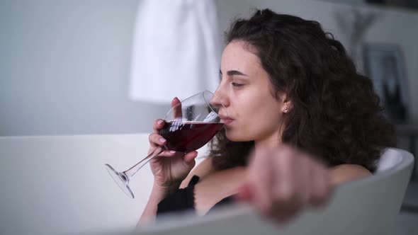 Young Brunette Woman Drinking Red Wine in Bathtub. Side View Close-up of Confident Caucasian Girl