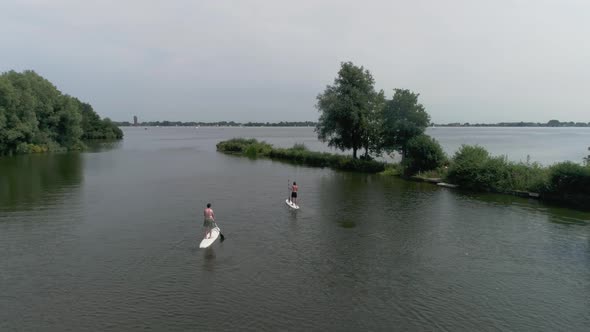 Drone Slomo shot of Two Male Friends Paddling on Surfboard towards a Lake, surrounded by Trees