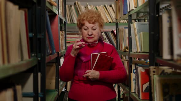 Smart Elderly Teacher in Glasses Holding Books in Her Hands in the Library