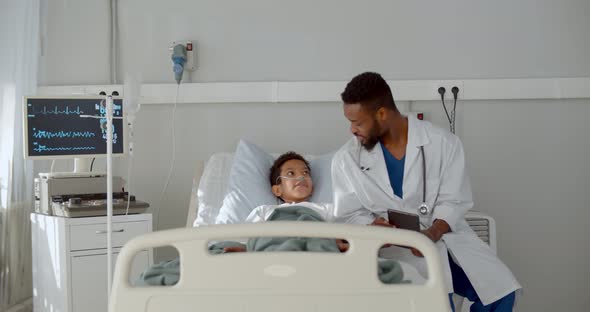 Young African Doctor Sitting on Hospital Bed and Talking To Preteen Boy Patient