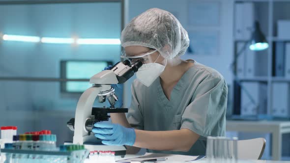 Female Lab Worker in Protective Uniform Looking through Microscope
