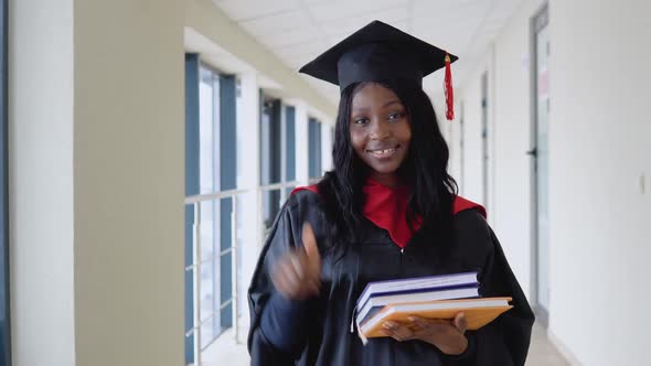 African American Female Graduate in Mantle Stands with a Books in Her Hands and Smiles