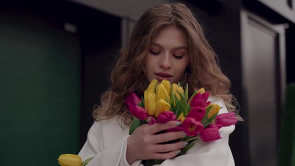 Nice Girl in a White Coat with a Bouquet of Tulips Looks at the Camera