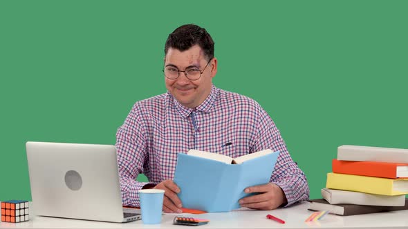 A Man with Glasses Sits at a Desk in Front of a Laptop and Reads a Book Smiles Puts His Fist to His