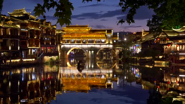 Fenghuang, China. Colorful Reflections of the Evening Town in the River. A Boat Is Sailing the River