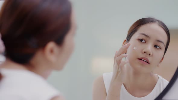 Fresh healthy skin of Asian girl looking at mirror, applying facial moisturizer on her face.