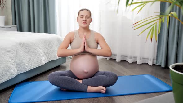 Beautiful Smiling Pregnant Woman Practicing Yoga Sitting on Fitness Mad in Lotus Pose and Breathing