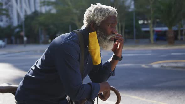 African american senior man wearing face mask talking on smartphone while leaning on his bicycle on