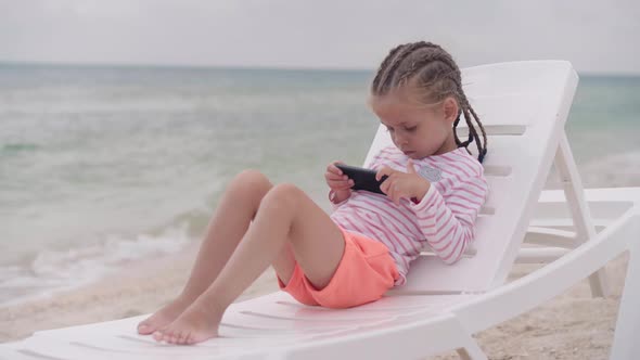 Little Girl Sitting Chaise Long Sea Beach Using Smartphone Watching Video Child is Bored