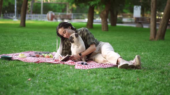 Cheerful Girl Laying on Plaid on Lawn in a Park and Making Notes While Little Pug Sitting Next to