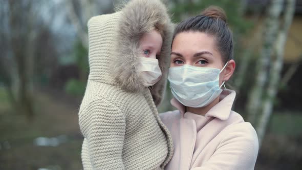 Mom with a small daughter in her arms, both in medical masks on a cool evening