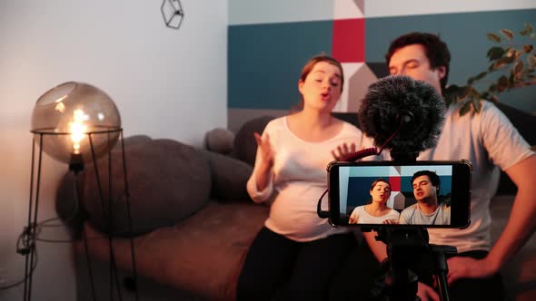 Man and Pregnant Woman Give an Interview Sitting on a Sofa in Living Room in Front of Mobile Camera