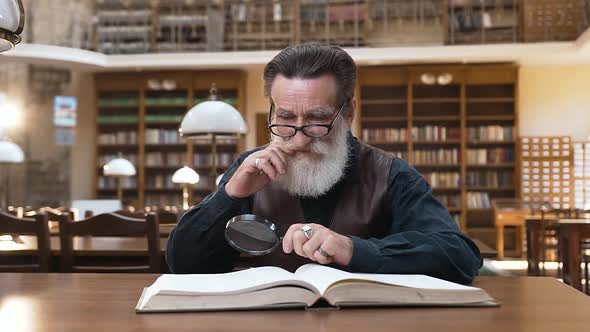 Old Intelligent Bearded Man in Glasses which Sitting in the Library and Reading Book