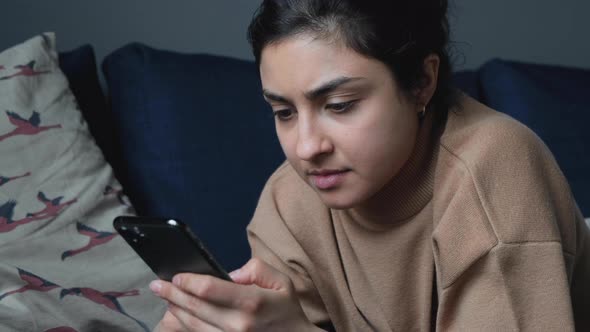 Close Up Of A Young Indian Woman Lying On A Bed Using A Smartphone Reading The News