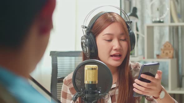 Young Asian Woman Holding Phone And Wearing Headphones Singing While Recording Podcast With Male