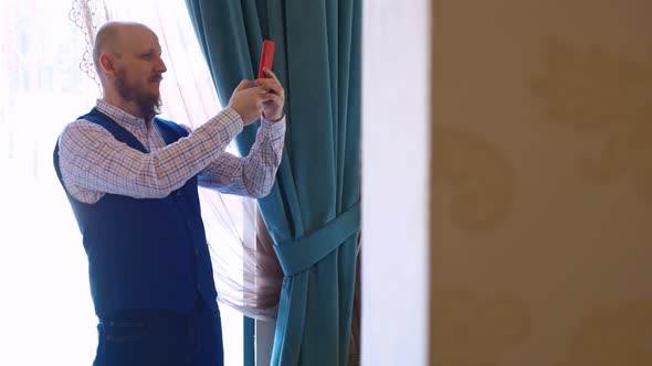 A Wellgroomed Man with a Beard in Classic Clothes Takes a Selfie While Standing By the Window