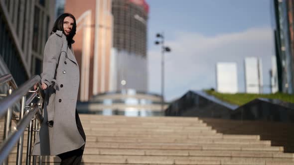 a Brunette in a Gray Coat Leans on the Railing and Stands on the Stairs Against the Backdrop of a