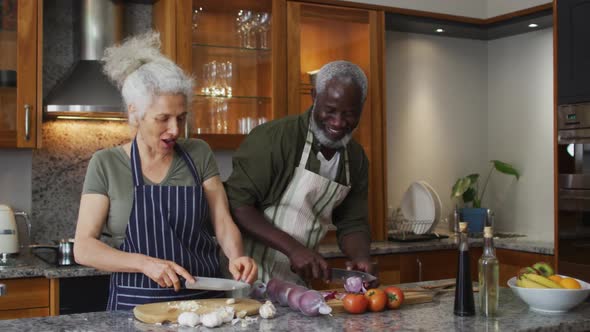 Sick mixed race senior couple wearing aprons chopping vegetables together in the kitchen at home